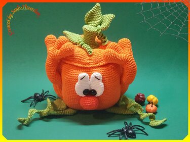 Gehäkelter oranger Kürbis mit weißen Augen, grünen Blättern und kleinen Dekokürbissen vor grünem Hintergrund