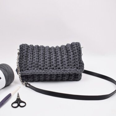 Dunkelgraue gehäkelte Clutch aus dickem Textilgarn mit Metallkette, schwarzem Lederriemen, daneben Garn, Häkelnadel und Schere auf weißem Hintergrund