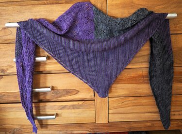 Mehrfarbiges gestricktes Dreieckstuch in Lila und Grau mit Loch- und Rippenmustern, drapiert an einer Stange vor einer Holzkommode.