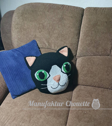 Schwarzes gehäkeltes Katzenkissen mit grünen Augen auf braunem Sofa.