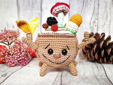 Nahaufnahme eines gehäkelten Waldgeist-Amigurumis mit Pilzen, roten und schwarzen Beeren und gelben Blättern auf dem Kopf vor Holzoberfläche und Naturdeko.