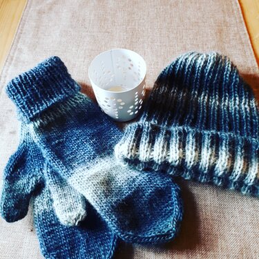 Gestrickte Mütze und Fäustlinge mit Farbverlauf in Blau- und Grautönen neben einem weißen Teelichthalter
