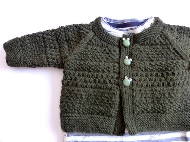 Dunkelgrüne, gestrickte Babyjacke mit drei hellgrünen Froschknöpfen über einem blau-weiß gestreiften Shirt auf weißem Hintergrund.