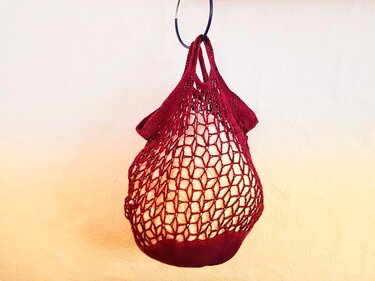 Bordeauxrote gehäkelte Netztasche mit offenem Maschenmuster, hängend an einem Haken vor einer hellen Wand
