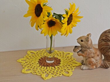 Gelb-braunes rundes Häkeldeckchen im Sonnenblumen-Look mit einer Vase voller Sonnenblumen und einer Eichhörnchenfigur.