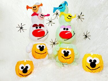 Zwei gestapelte gehäkelte Amigurumi-Figuren (weiß und hellgrün) mit roten Nasen, vorn je ein gehäkelter Kürbis und kleinen bunten Geistchen auf den Köpfen; schwarze Spinnen-Deko auf weißem Hintergrund