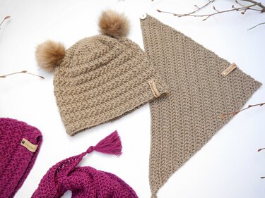 Beige crocheted baby beanie with two faux-fur pom-poms next to a beige triangular crochet bandana with a button; magenta crochet pieces partially visible