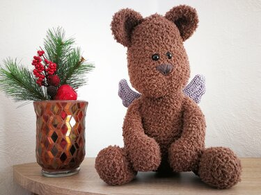Brauner Amigurumi-Teddybär mit lila Engelsflügeln neben Tannengrün und roten Beeren.