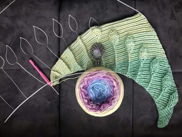 Halbfertiges gehäkeltes Dreieckstuch mit reliefartigen Blattmotiven, mehrfarbigem Farbverlaufsgarn und Häkelnadel auf dunklem Untergrund