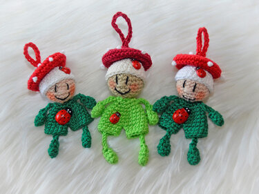 Drei gehäkelte Glückspilz-Anhänger mit roten Pilzhüten und grünen Körpern, verziert mit Marienkäfer-Buttons auf hellem Untergrund.