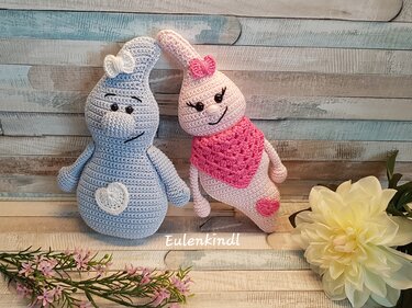 Zwei gehäkelte Amigurumi in Pastellblau und Rosa mit Herzapplikationen und Schleifen vor Holzbrett