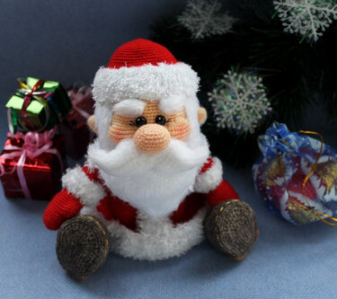 Gehäkelte Amigurumi-Weihnachtsmann-Puppe in Rot und Weiß, sitzend vor Weihnachtsdekoration und Geschenken.