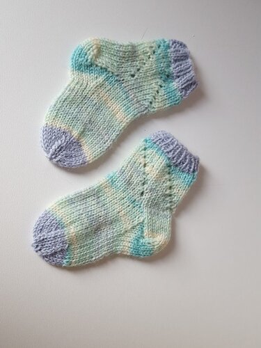 Zwei kleine gestrickte Babysocken in Pastellfarben (Blau, Creme, Lila) mit dezentem Lochmuster.