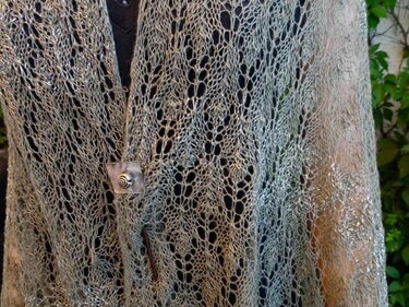 Nahaufnahme eines hellgrauen Lacetuchs mit offenem Lochmuster und quadratischer Brosche über dunkler Kleidung