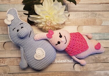 Zwei Amigurumi-Häkelpuppen: links hellblau mit Herz, rechts rosa mit Schleife und Schultertuch, auf hellem Holzbrett liegend