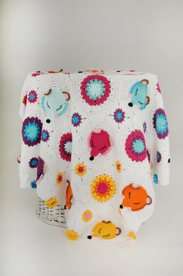 White crochet blanket with colorful sleeping fox and flower appliques draped over a white wicker basket.
