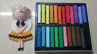 Gehäkelter Engels-Amigurumi mit gelbem Kleid, weißen Flügeln und Schleife neben farbigen Pastellkreiden