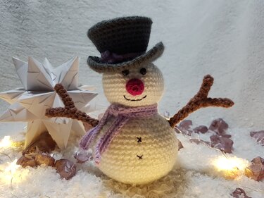 Gehäkelter Schneemann mit Zylinder und lila Schal, umgeben von Kunstschnee, Origami-Stern und Lichterkette.