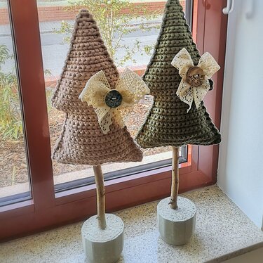 Zwei gehäkelte Tannenbäume in Beige und Olivgrün mit Spitzen-Schleifen und Zierknöpfen auf Holzstäben in grauen Sockeln vor einem Fenster.