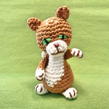 Braun-weißes gehäkeltes Amigurumi-Kätzchen mit grünen, gestickten Augen auf grünem Hintergrund