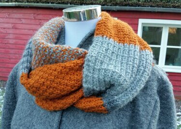 Dicker, gestrickter Loop-Schal in Grau und Orange auf einer Büste vor einem roten Holzhaus.