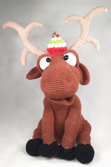 Brown crocheted amigurumi moose plush toy wearing a small red, white, and green Christmas hat with antlers.