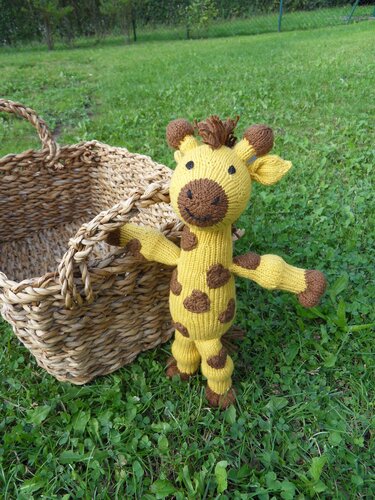 Gestrickte gelbe Amigurumi-Giraffe mit braunen Flecken im Gras neben einem geflochtenen Korb.