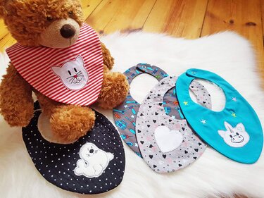 Fünf Baby-Lätzchen mit verschiedenen Applikationen (Katze, Robbe, Einhorn, Herz, Werkzeugmotive) auf hellem Untergrund; ein Teddybär trägt ein gestreiftes Lätzchen.