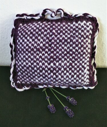 Gestricktes quadratisches Dekoelement in Lila und Weiß mit gedrehtem Rand und Schlaufe, davor Lavendelstiele