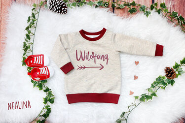 Beiges Baby‑Sweatshirt mit dunkelrotem Bündchen und Plottmotiv ‚Wildfang‘ mit Pfeil, arrangiert mit roten Babyschuhen und Efeuranken auf hellem Felluntergrund.