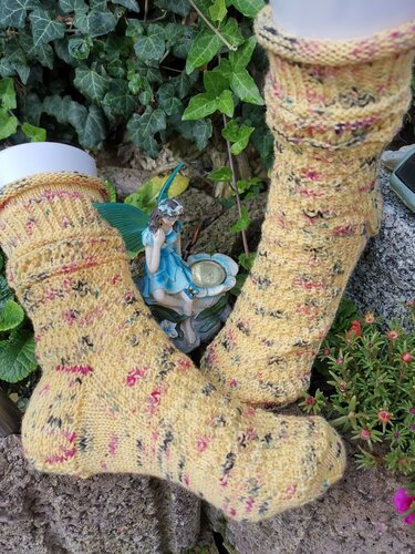 Handgestrickte gelbe Socken mit bunten Sprenkeln auf weißen Sockenhaltern, auf Steinen im Garten neben einer kleinen Feenfigur und Efeu platziert.