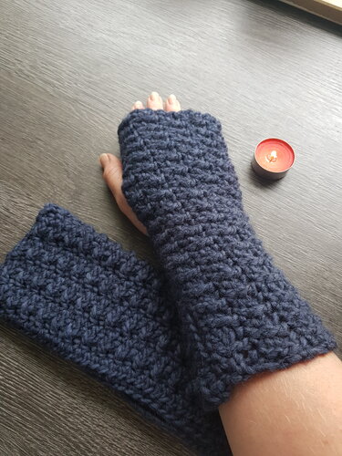 Dunkelblaue gehäkelte fingerlose Stulpe am Unterarm, zweite Stulpe liegt auf dunklem Holztisch neben einem Teelicht