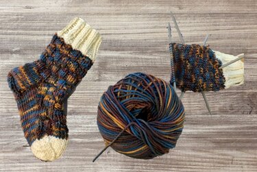 Handgestrickte Kindersocke mit Wollknäuel und Stricknadeln, angefangene Socke auf Holzuntergrund.