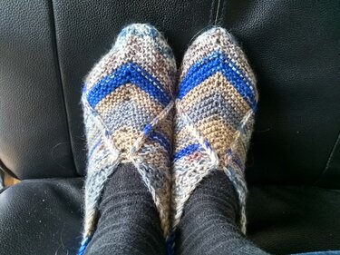 Zwei handgestrickte Hausschuhe in Grau, Beige und Blau mit Zickzackmuster, getragen über dunklen Socken auf schwarzem Sitz.
