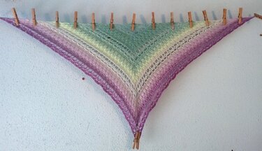 Dreieckiges gehäkeltes Tuch in Lila‑Grün mit Spitzen‑ und Lochmuster, an Wäscheklammern an der Wand gespannt