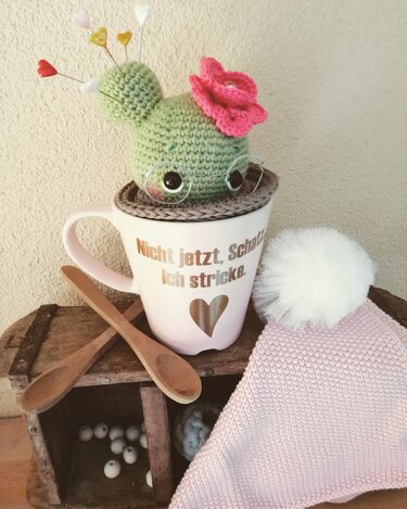 Gehäkelter grüner Kaktus mit rosa Blüte in einer Tasse als Nadelkissen, herzförmige Stecknadeln, Holzlöffel und rosafarbene Strickmütze