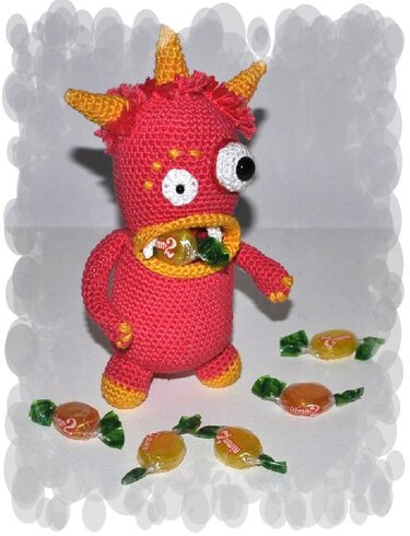 Gehäkeltes rotes Monster mit gelben Hörnern, großen Augen und Bonbon im Mund; verpackte Bonbons auf dem Boden