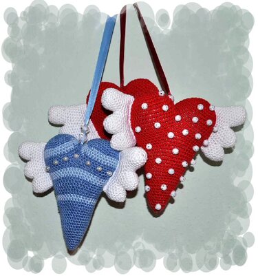 Zwei gehäkelte Amigurumi-Herzen (rot und blau) mit weißen Flügeln und Perlen, an Satinbändern hängend