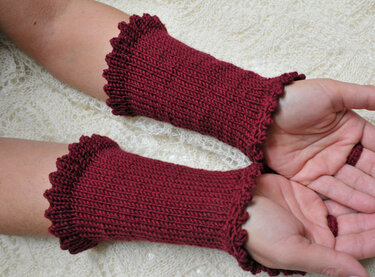 Weinrote, gestrickte Armstulpen mit Rüschensaum an den Handgelenken einer Person vor hellem Hintergrund.