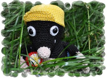 Schwarzer gehäkelter Maulwurf mit gelber Mütze und rosa Schnauze, sitzt im Gras und hält ein kleines rotes Objekt.