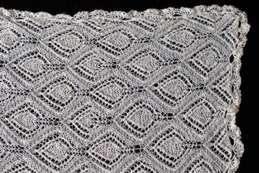 Weißes gestricktes Lacetuch mit Rautenmuster und gewellter Spitzenkante auf schwarzem Hintergrund.