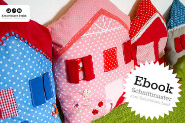 Drei dekorative Hauskissen in Rosa, Blau und Rot mit applizierten Fenstern, Türen und Blumenmotiven.