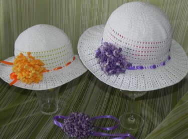 Erwachsenen- und Kinder-Sonnenhut aus weißem Häkelstoff mit eingeflochtenen Bändern und separater lila Blumenapplikation auf grünem Hintergrund