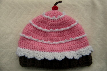 Gehäkelte Babymütze im Cupcake‑Stil mit braunem Bund, rosa Oberteil, weißen Rüschen und roter Kirsche