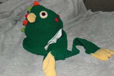 Grüne gehäkelte Babymütze im Drachen-Design mit Augen, bunten Stacheln und langen Ohrenklappen mit gelben Enden