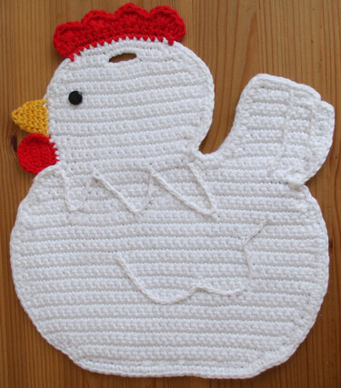 Weißer gehäkelter Topflappen in Huhnform mit rotem Kamm, gelbem Schnabel und Knopfauge auf Holzuntergrund.