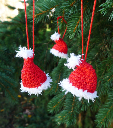 Drei gehäkelte rote Weihnachtsmützen mit weißem Rand an Tannenzweigen hängend