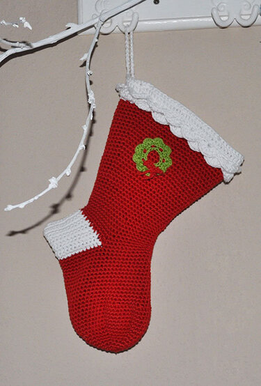 Gehäkelter roter Weihnachtsstrumpf mit weißem Bündchen und grünem Kranz, an einem Haken hängend.