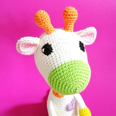 Nahaufnahme einer gehäkelten Amigurumi-Giraffe in Weiß mit grünem Maul, orangen Hörnern und schwarzen Augen vor pinkem Hintergrund.