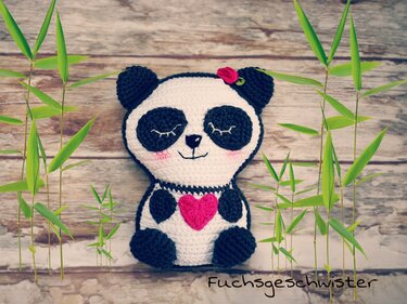 Gehäkeltes Panda-Amigurumi mit rosa Herz, Blüte am Ohr und geschlossenen Augen vor Holzuntergrund und Bambus